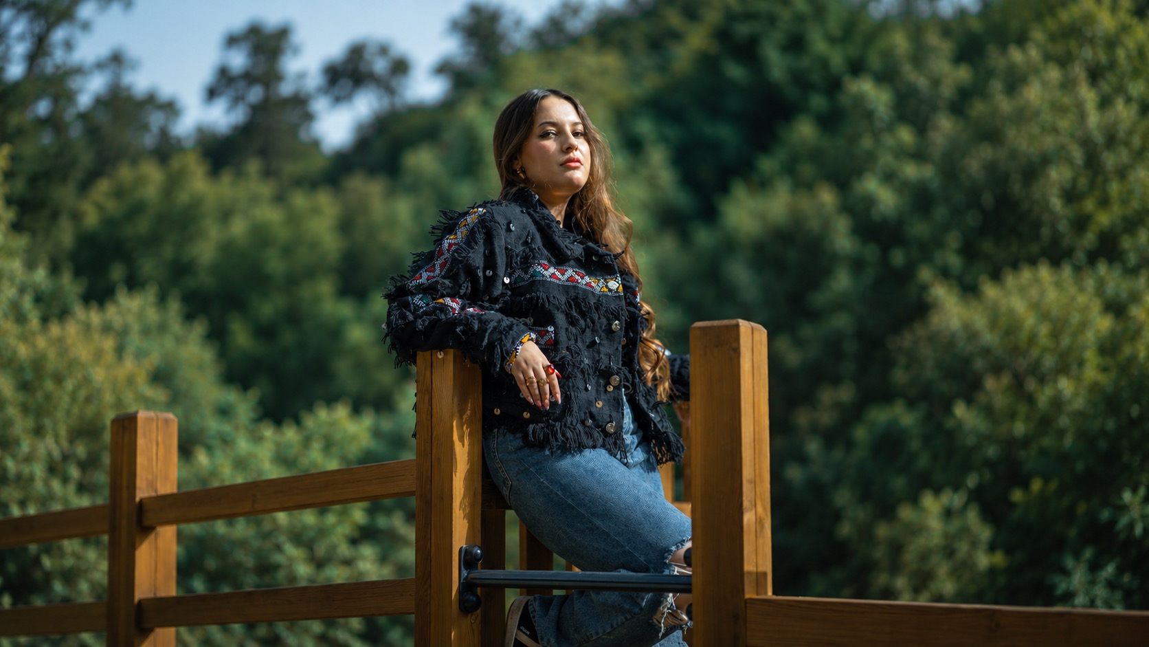 Model in a black embellished jacket leaning on a wooden railing in front of a forest